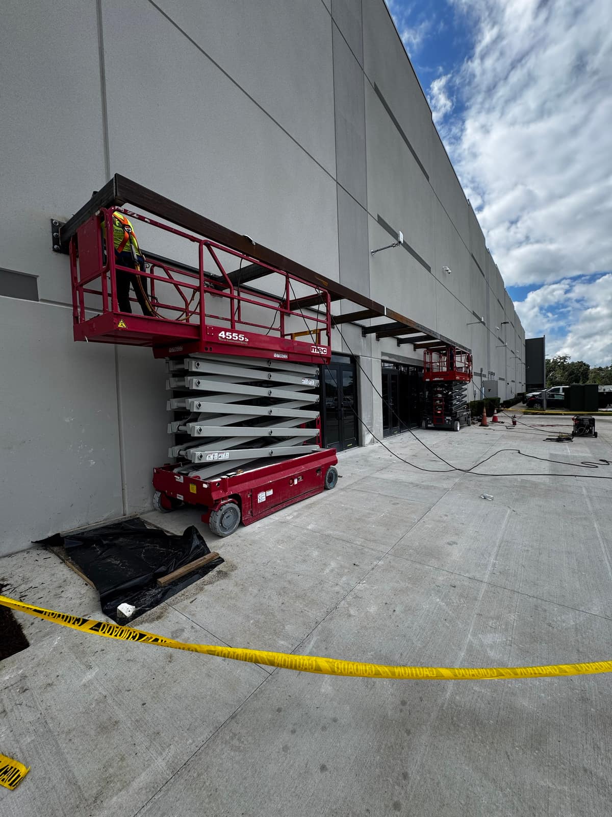 The canopy steel running along the building facade past several entrances, with two scissor lifts and caution tape below