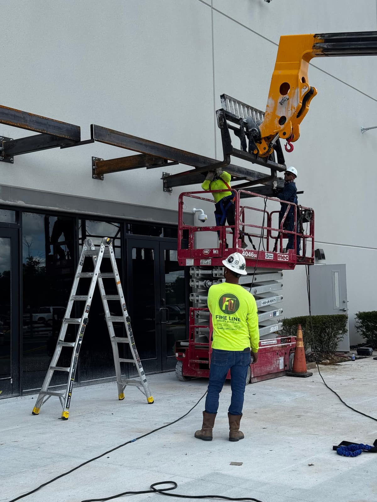 Telehandler holding a long steel tube while two crew members on a scissor lift guide it onto the cantilever brackets, with a Fine Line Weld & Build crew member watching from the ground