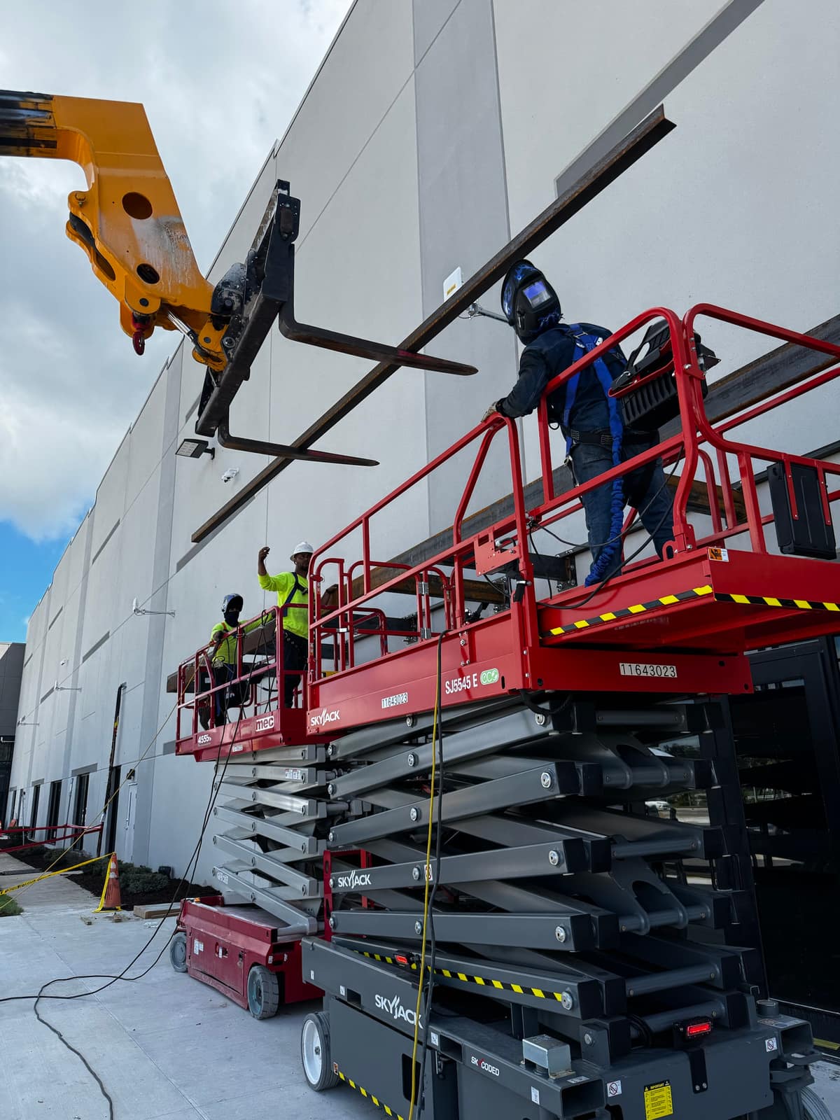 Welder in a hood and harness working from a scissor lift while a telehandler holds the beam against the tilt-wall, with two more crew on a second lift