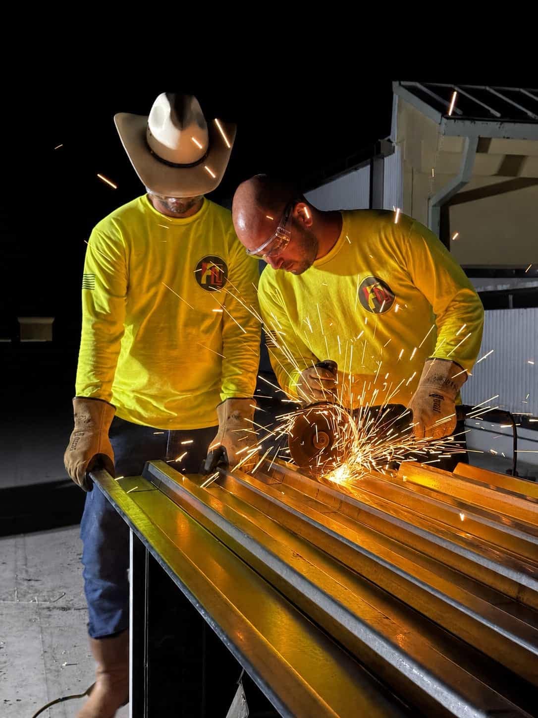 Two Fine Line Weld & Build crew members in branded hi-vis shirts cutting a metal deck panel with an angle grinder, sparks flying across the sheet