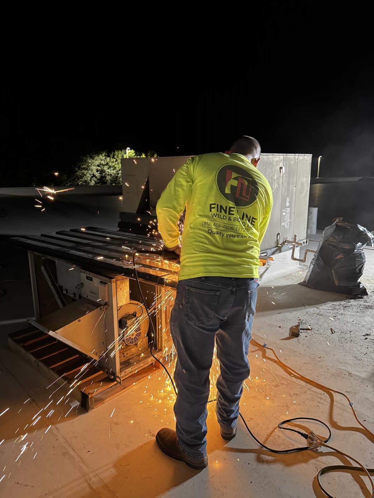 Fine Line Weld & Build crew member cutting steel with an angle grinder on the roof after dark, sparks spraying across the surface beside a rooftop HVAC unit
