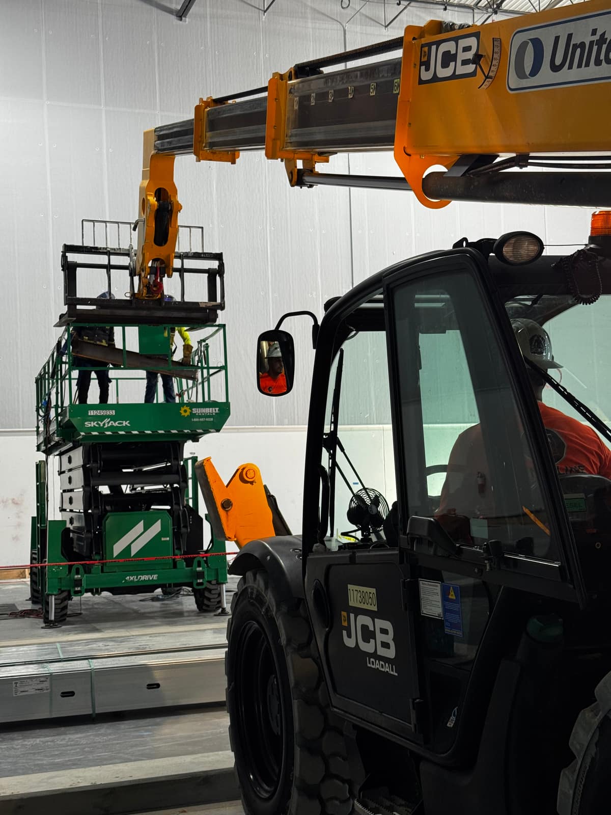 Workers on a scissor lift handling a steel member while a telehandler boom holds material overhead inside a warehouse