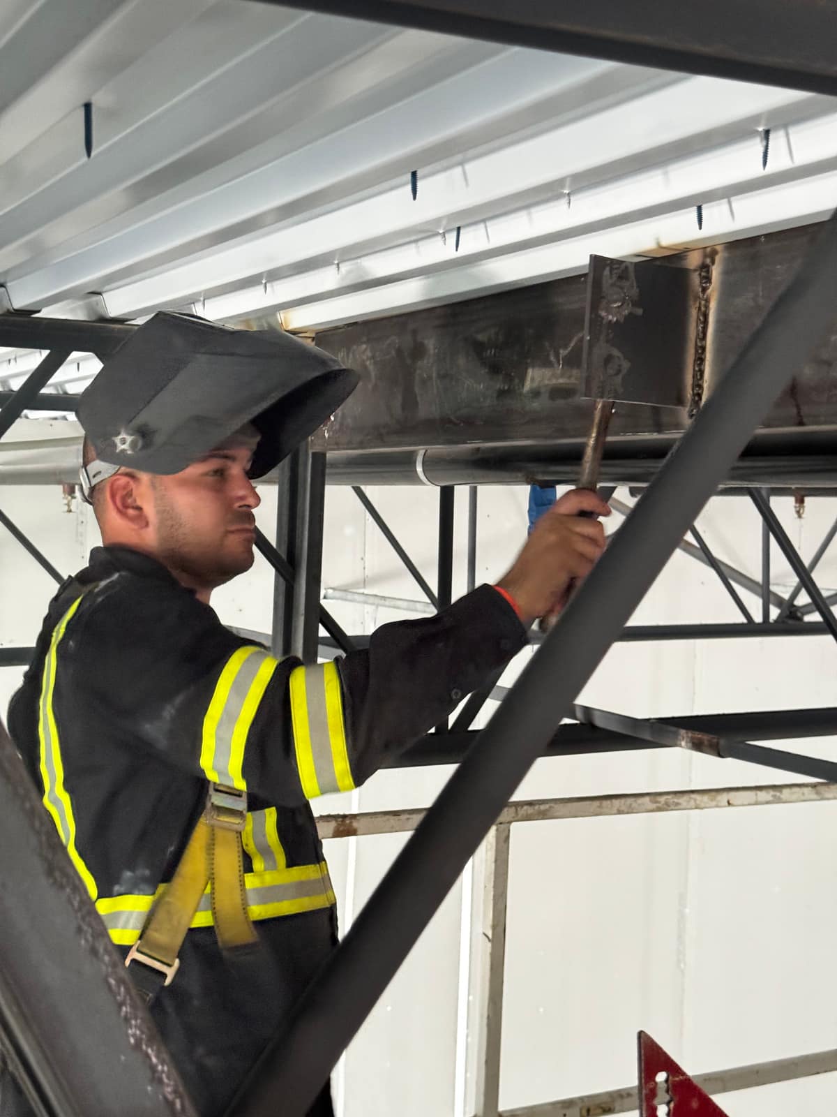 Welder in flame-resistant gear checking a finished weld on a plate connection below the roof deck