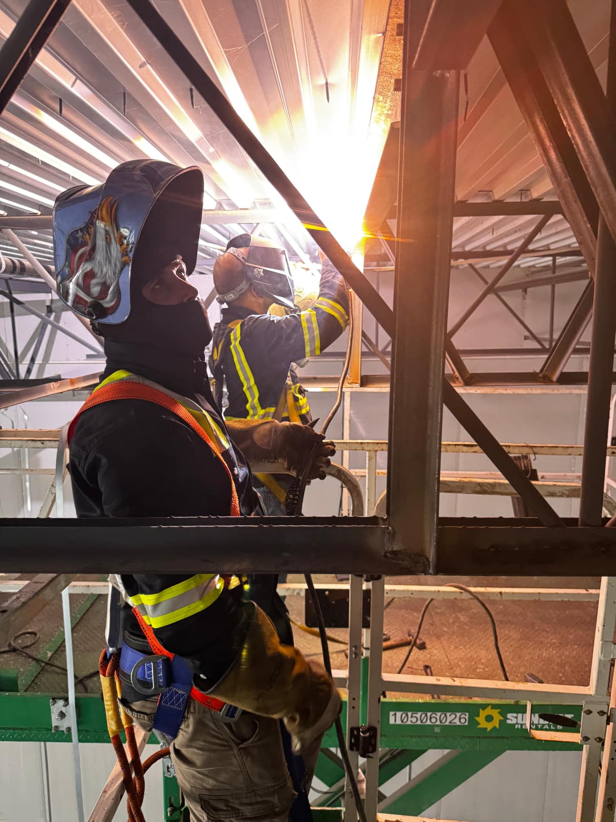 Two welders in harnesses working from a scissor lift at roof level, arc flaring against the steel