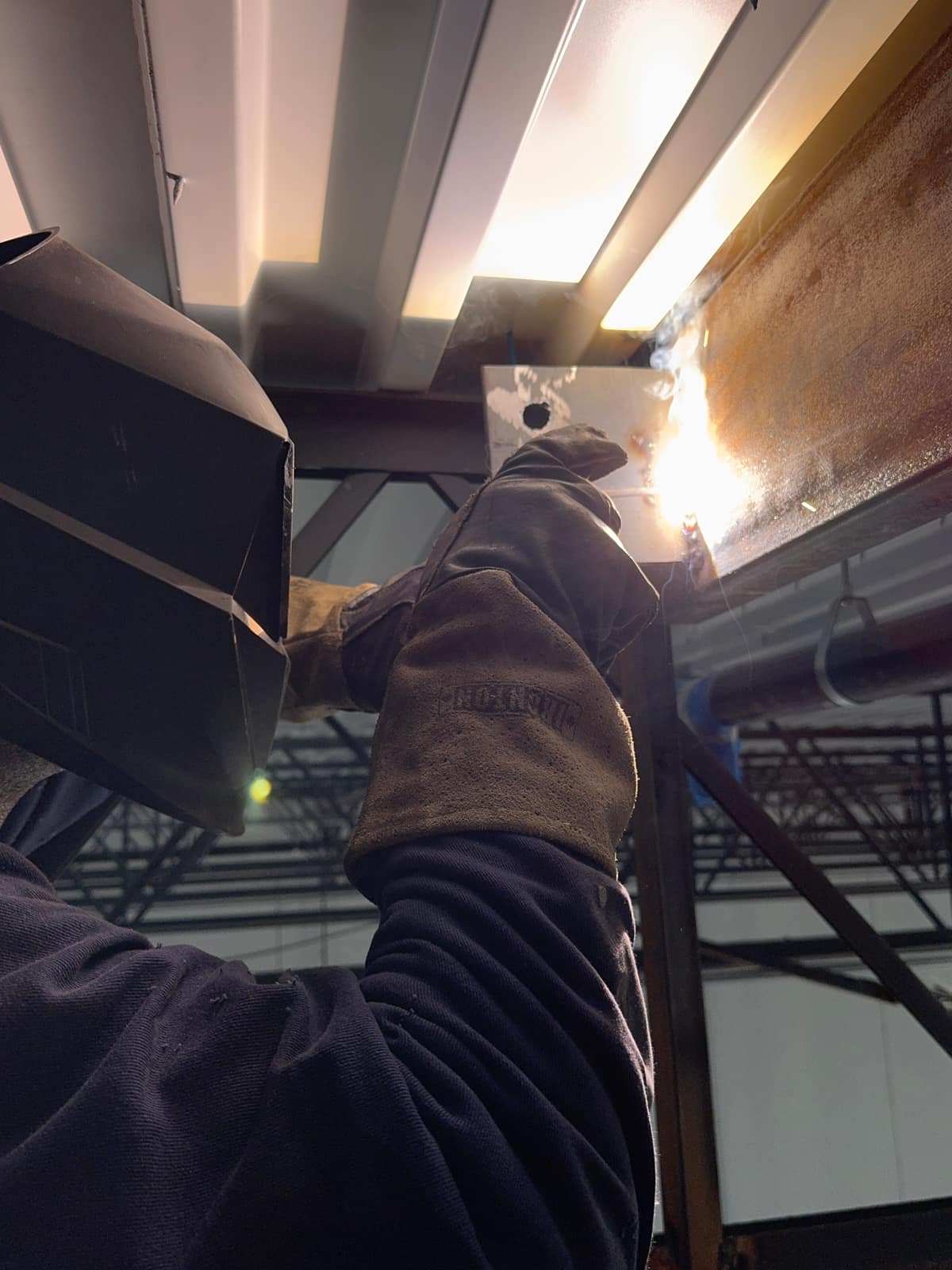 Welder looking up behind a hand-held hood, running an overhead weld on a steel connection plate at the beam above