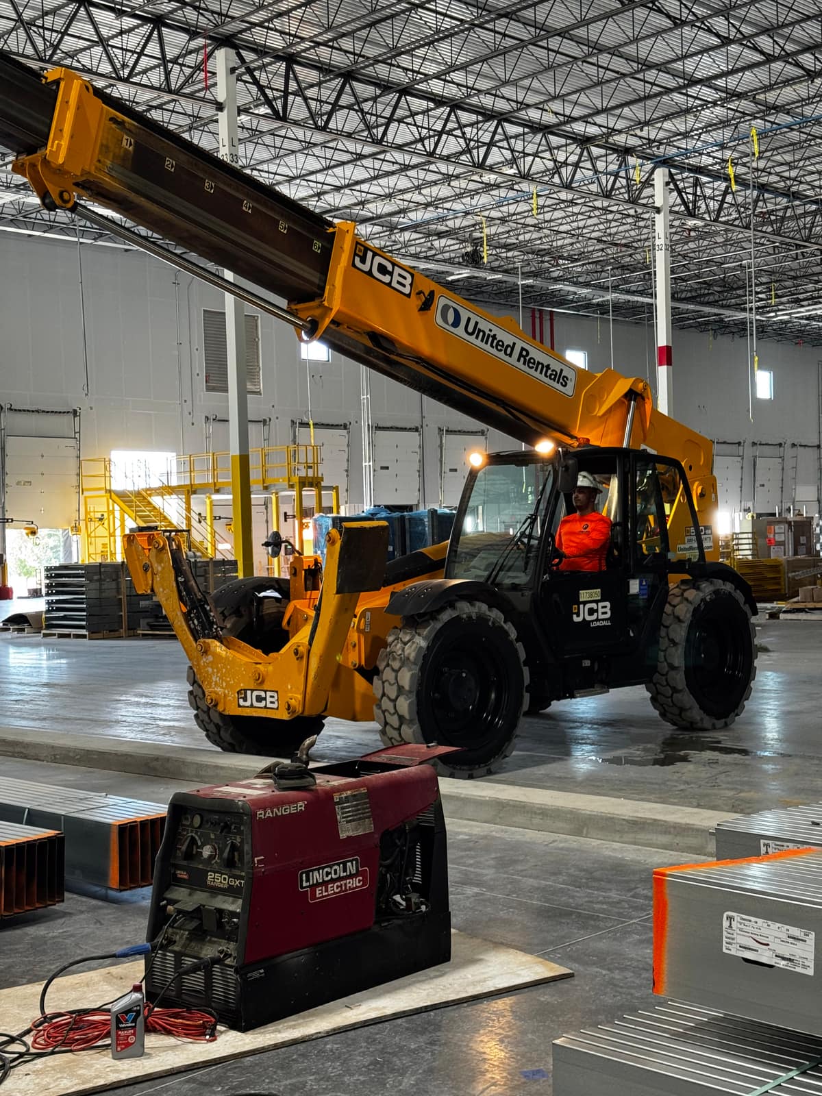 JCB telehandler with its boom raised inside a warehouse, with an engine-driven Lincoln Electric welder on the concrete floor in front