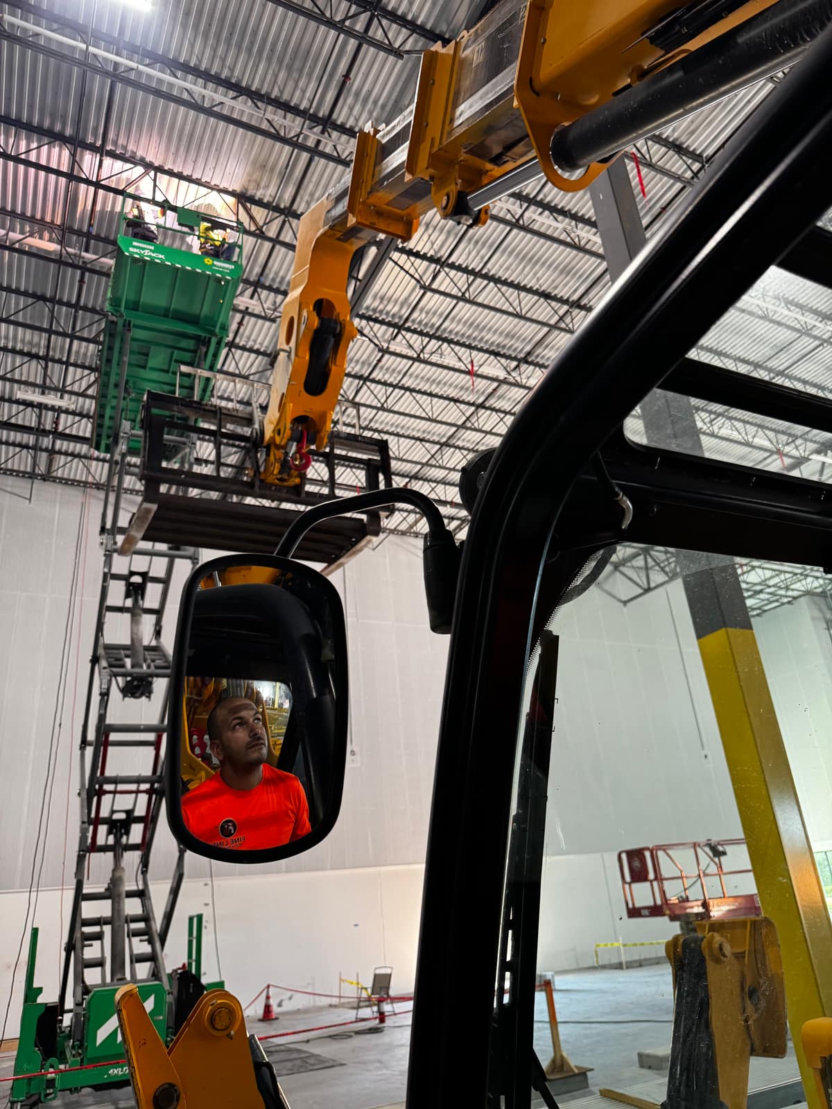 View from inside the telehandler cab toward the warehouse roof structure, with the operator reflected in the mirror